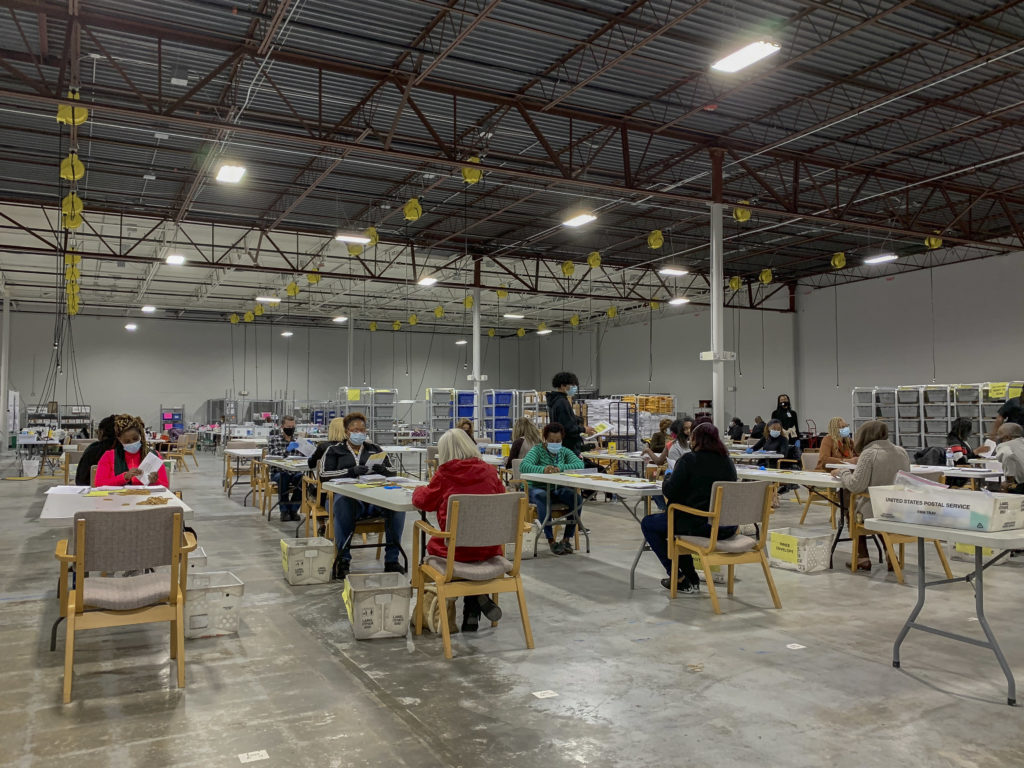 Photo of several pairs of people at opposite ends of folding conference tables, handling piles of paper. People are a variety of ages, all wearing COVID masks. Most appear to be women, both Black and white. Room has concrete floors, visible vertical supports and ceiling beams. Carts with stacked plastic baskets and other equipment are in background. Observers can help protect the vote count.