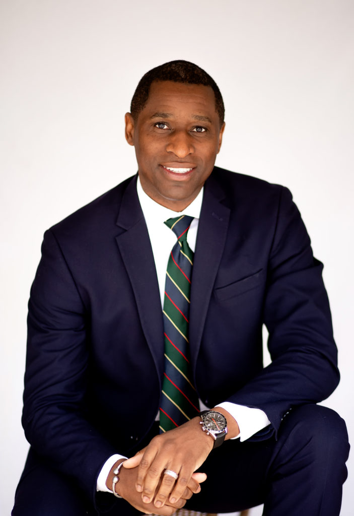 Portrait of Bennie Smith, a Black man in a blue suit and tie, with short hair and his hands clasped, one elbow resting on bent knee.