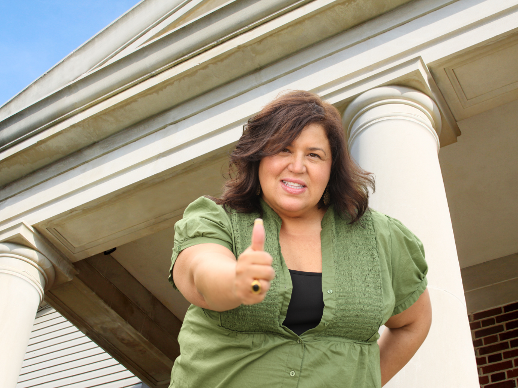 Casually dressed brown-skinned woman showing "thumbs up" sign, standing in front of government building. Poll tapes provide a method of citizen oversight of elections.