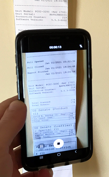 Phone held upright by light-skinned person's left hand, with screen showing a poll tape being video-recorded. Poll tape can be seen behind phone and appears to be hanging on a wall.